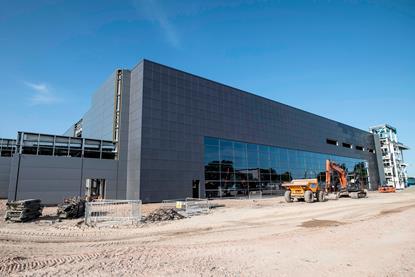 A photo of a grey blocky building with a large glass front under a bright blue skye. The building is surrounded by construction sand, there is a digger standing in front of the building and the top of a crane is poking over the top of the roof.