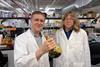 Two scientists in lab coats in a modern lab. One is holding a glass flask with a dark goo inside.