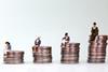 Models of tiny people sitting on stacks of coins. A man is on the tallest stack and a mother and baby is on the smallest with two other woman in between.