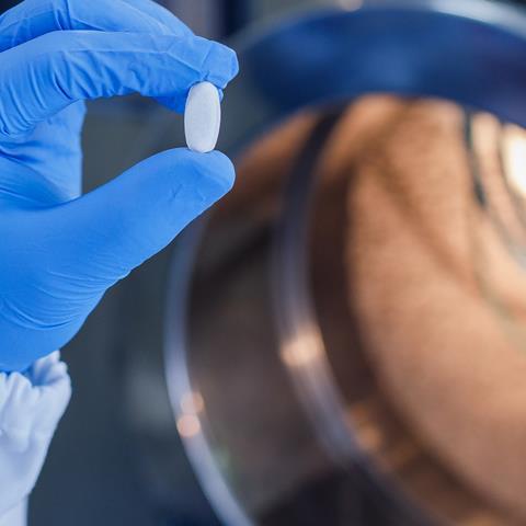 A gloved hand holds a pill up to the camera in a pharmaceutical lab