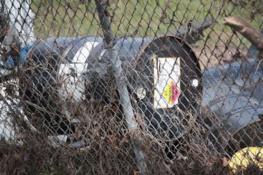 An oil drum that has been moved by bad weather and a flood resting against a chain link fence